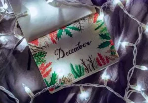 A high-angle close-up photograph of an open notebook or journal, showing a page decorated with a hand-painted border of holly, red berries, and other winter greenery. The word "December" is written in elegant script in the center of the page. The notebook is resting on a dark, textured surface and is surrounded by a string of glowing white LED Christmas lights.