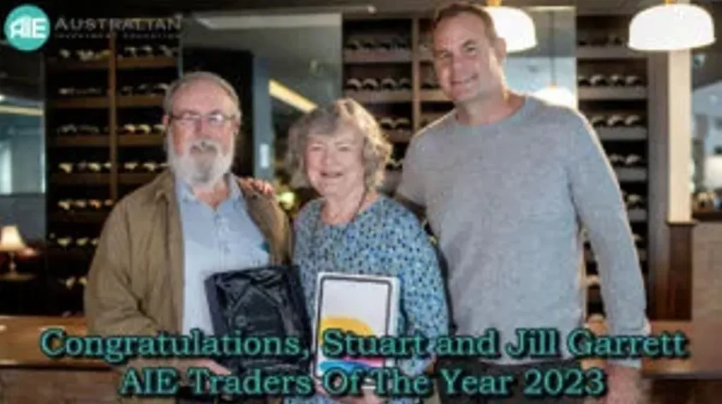 A photograph of three people: an older man with a beard holding a dark glass award plaque, an older woman holding a white tablet, and a younger man standing to the right. They are all smiling in what appears to be a room with wine racks in the background. Text overlay on the image reads: "Congratulations, Stuart and Jill Garrett AIE Traders Of The Year 2023."