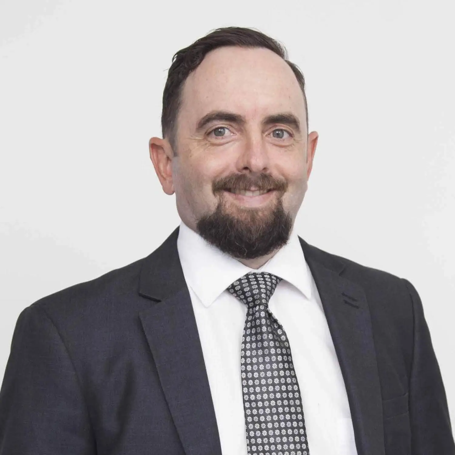 Professional headshot of a smiling man, Andrew Gibbs, with a beard, wearing a dark suit and a patterned tie.