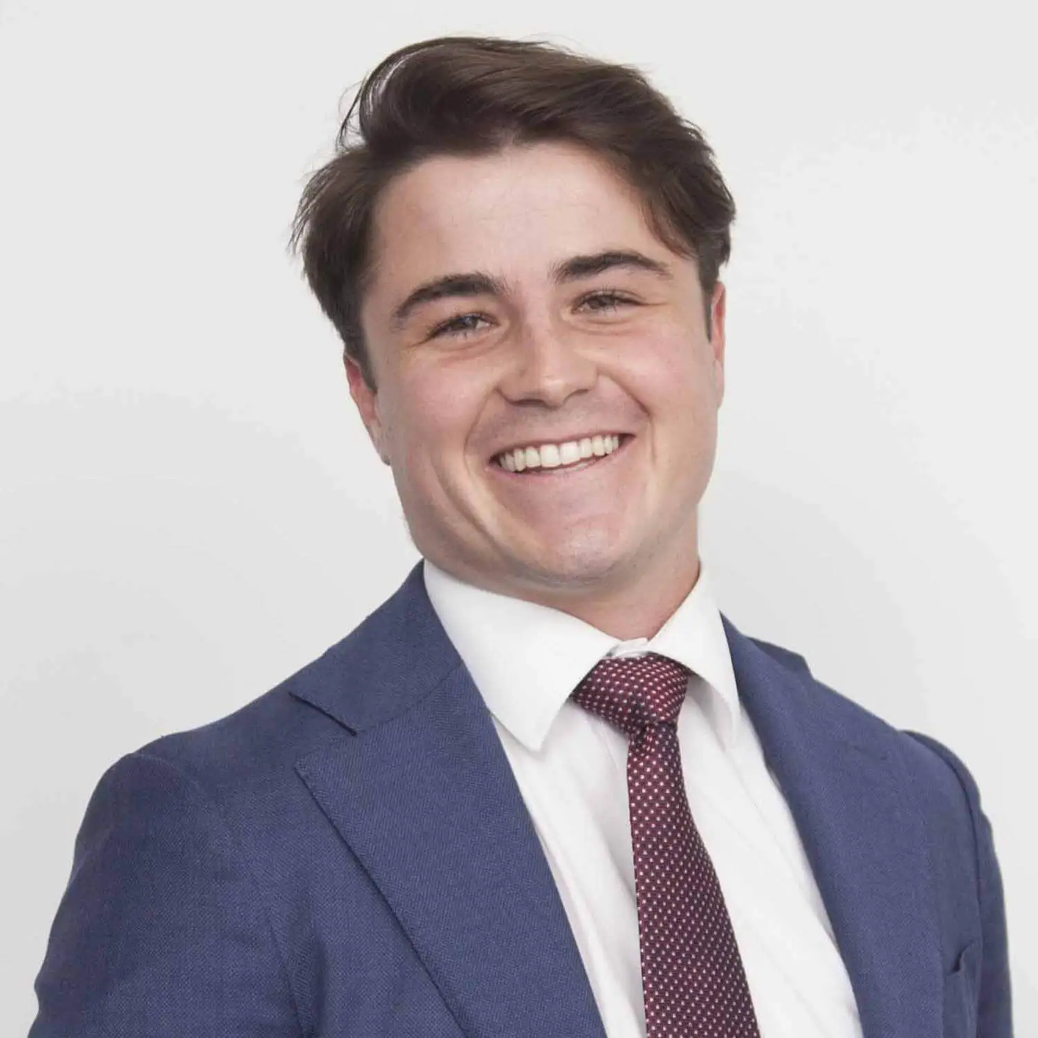 Professional headshot of a smiling young man named Lewis Ioannou in a blue suit and a patterned red tie.