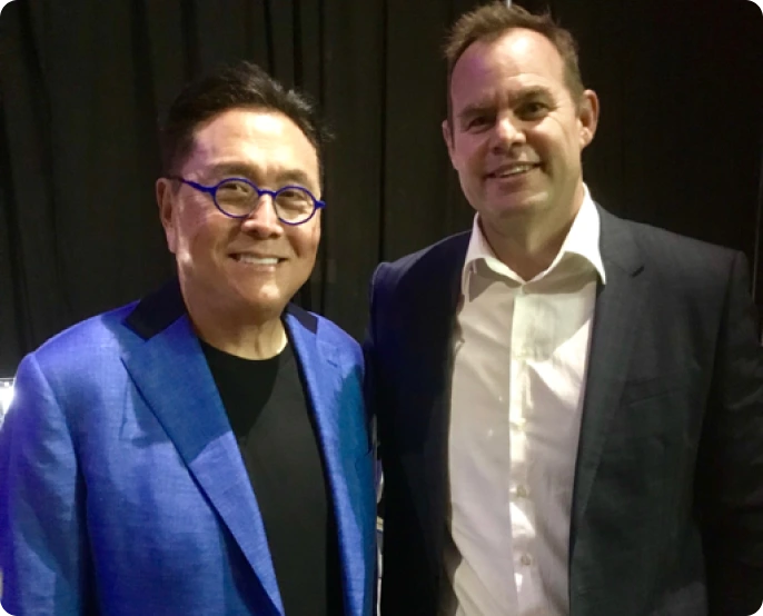 A photo of Robert Kiyosaki, author of Rich Dad Poor Dad, wearing a blue blazer and glasses, standing next to another man in a dark suit and white shirt.