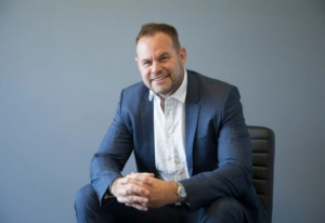 A smiling professional man with a beard sits in an office chair, wearing a blue plaid suit and an open-collar white shirt, with his hands clasped and a plain gray wall behind him.