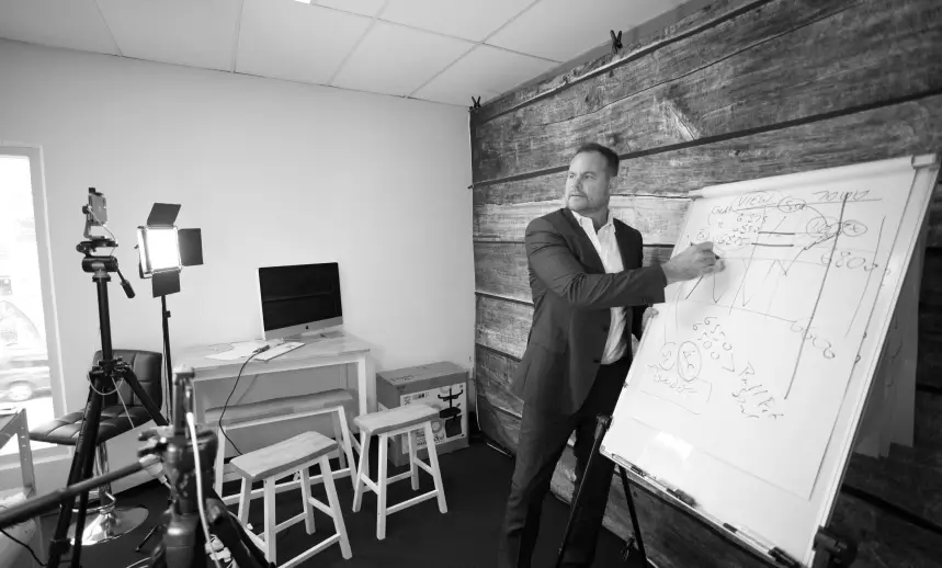 A black and white photo of a man in a suit drawing a complex chart on a whiteboard in a small studio with video lighting and a computer monitor visible.